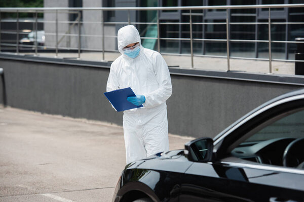 Medical worker in hazmat suit and goggles writing on clipboard near blurred car 