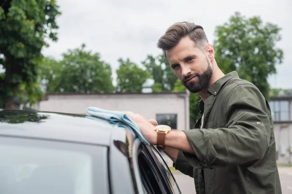 Bearded driver cleaning blurred car with rag outdoors