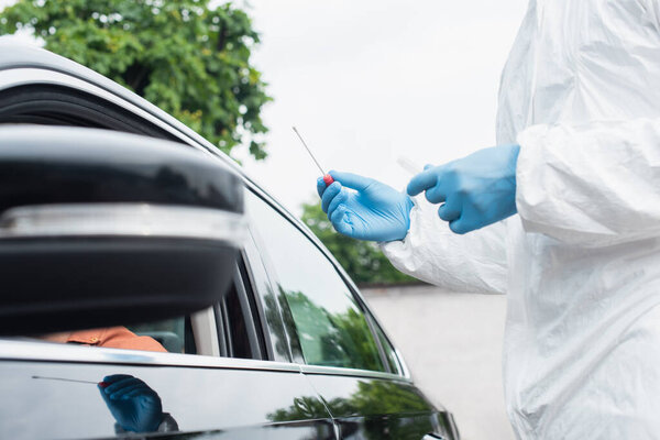 Cropped view of medical worker in hazmat suit holding pcr test near driver in blurred car
