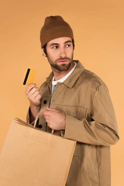 young man in beanie and jacket holding shopping bags and credit card isolated on beige
