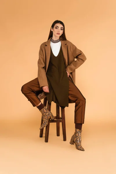 穿着时髦外套和靴子的年轻女子坐在米色背景的木制凳子上 — 图库照片