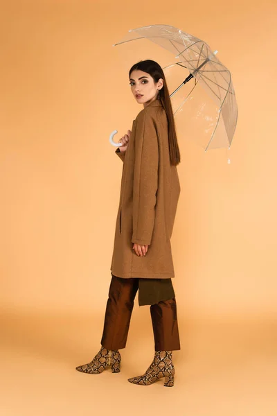 ベージュの背景に透明な傘の下に立つ茶色のコートと革のブーツのきれいな女性 — ストック写真