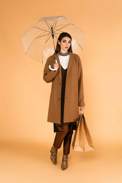 Modische Frau Steht Mit Einkaufstaschen Unter Transparentem Regenschirm Auf Beige — Stockfoto