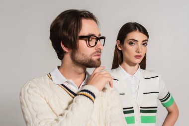 brunette woman in striped polo t-shirt looking at camera near man touching chin isolated on grey
