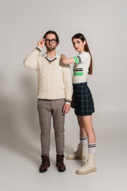 man in white pullover adjusting eyeglasses near woman leaning on his shoulder on grey background