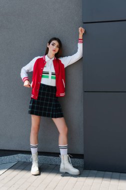 full length view of young woman in stylish clothes posing near grey wall outdoors