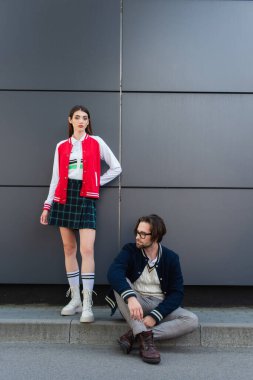 young stylish woman looking at camera near trendy man sitting on border outdoors