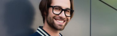 pleased man in trendy eyeglasses looking at camera near grey wall, banner