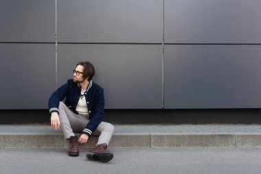 young man in trendy autumn clothes looking away while sitting on border near grey wall