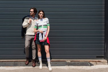 woman in striped polo t-shirt, plaid skirt and boots posing near trendy man and grey wall