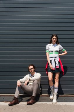 stylish woman standing with hand on hip near man sitting on border of urban street