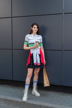 trendy woman with coffee to go and shopping bags smiling near grey wall 