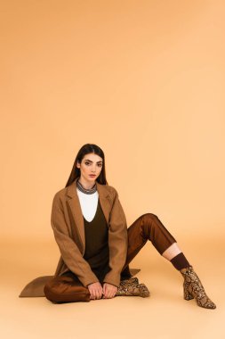 pretty woman in trendy coat, satin pants and leather boots sitting on beige background