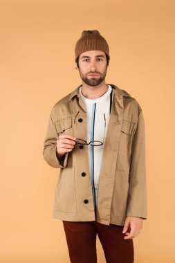 front view of young man in stylish jacket and beanie holding eyeglasses isolated on beige