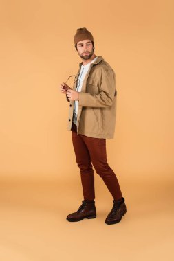full length view of trendy man looking back while holding eyeglasses on beige