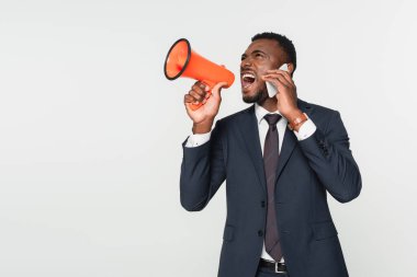 african american businessman in suit screaming in megaphone and talking on smartphone isolated on grey clipart