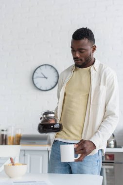 african american man holding coffee pot near cup in kitchen clipart