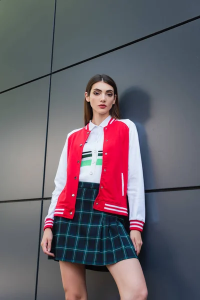 low angle view of brunette woman in stylish jacket and plaid skirt near grey wall