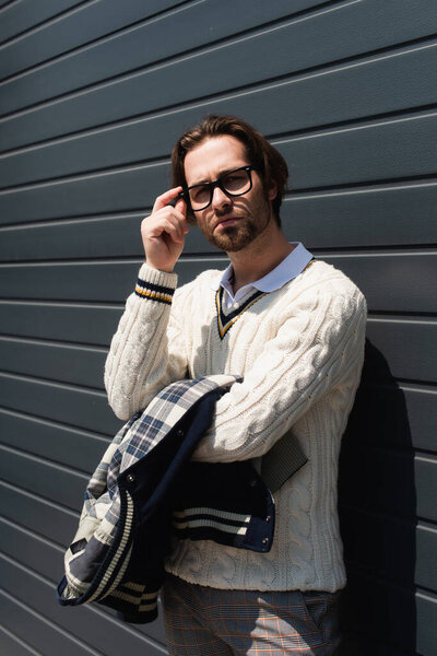 trendy man in white knitted sweater adjusting eyeglasses near grey wall outdoors