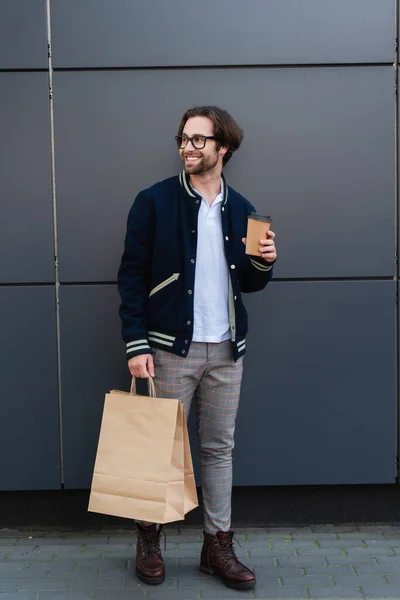 紙コップと買い物袋を持った灰色の壁のそばに立つ陽気でスタイリッシュな男 — ストック写真