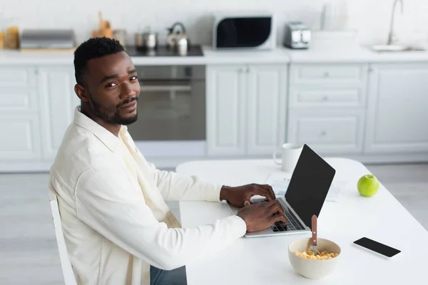 年轻的非洲裔美国自由职业者在厨房里使用智能手机旁边有空白屏幕的笔记本电脑 — 图库照片