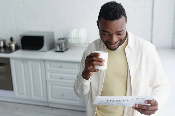 陽気なアフリカ系アメリカ人男性がコーヒーを持って旅行生活新聞を読んで — ストック写真