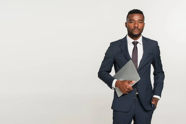 スーツを着たアフリカ系アメリカ人ビジネスマンがグレーで隔離されたポケットに手を置きながらノートパソコンを持ち — ストック写真