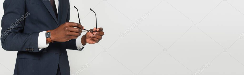 cropped view of african american man in suit holding eyeglasses isolated on grey, banner