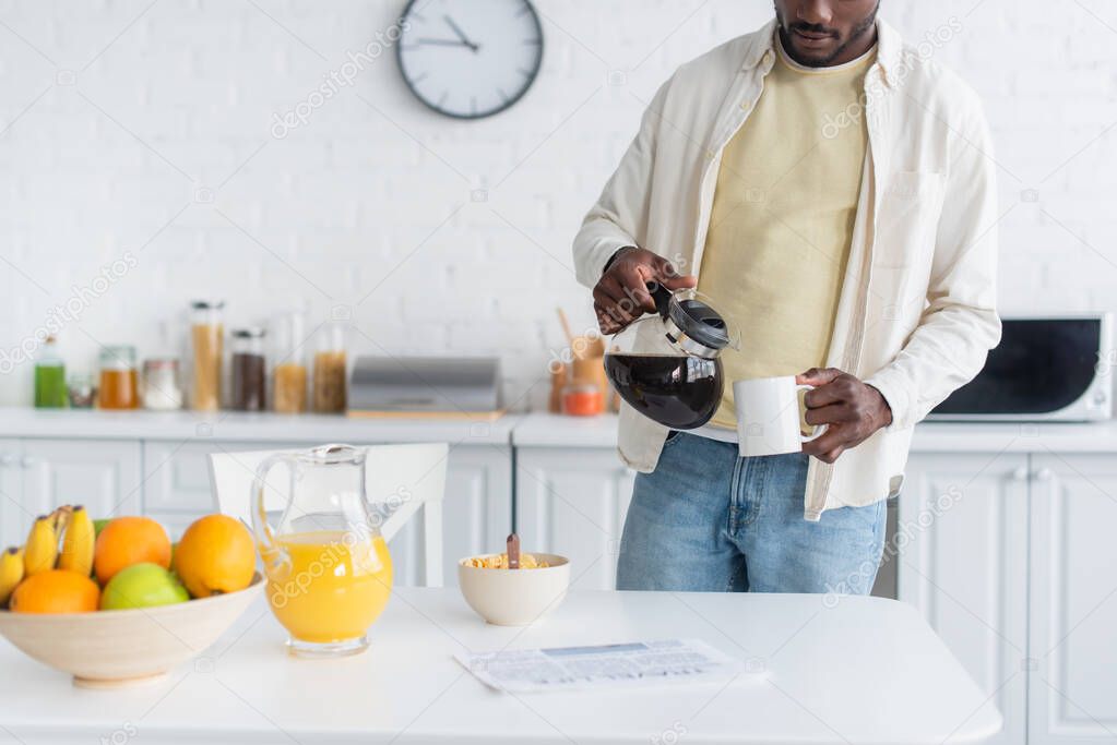 cropped view of bearded african american man holding coffee pot near cup in kitchen