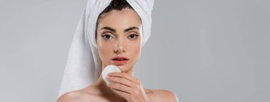 young woman with towel on head removing makeup with cotton pad isolated on grey, banner clipart