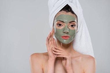 Kafasında havlu ve yeşil kil maskeli genç bir kadın gri renkli kameraya bakıyor.