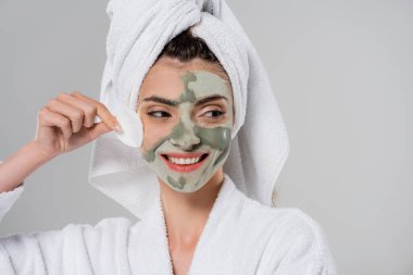 Bornozlu mutlu genç kadın, gri üzerine izole edilmiş pamuk yastıkla kilden maske çıkarıyor. 