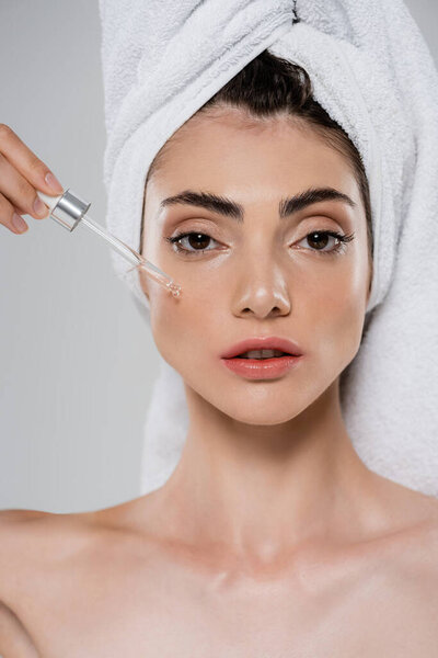 красивая молодая женщина с полотенцем на голове применяя витамин С сыворотку с пипеткой изолированы на сером