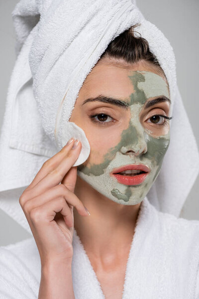 young woman in bathrobe removing clay mask with cotton pad isolated on grey 