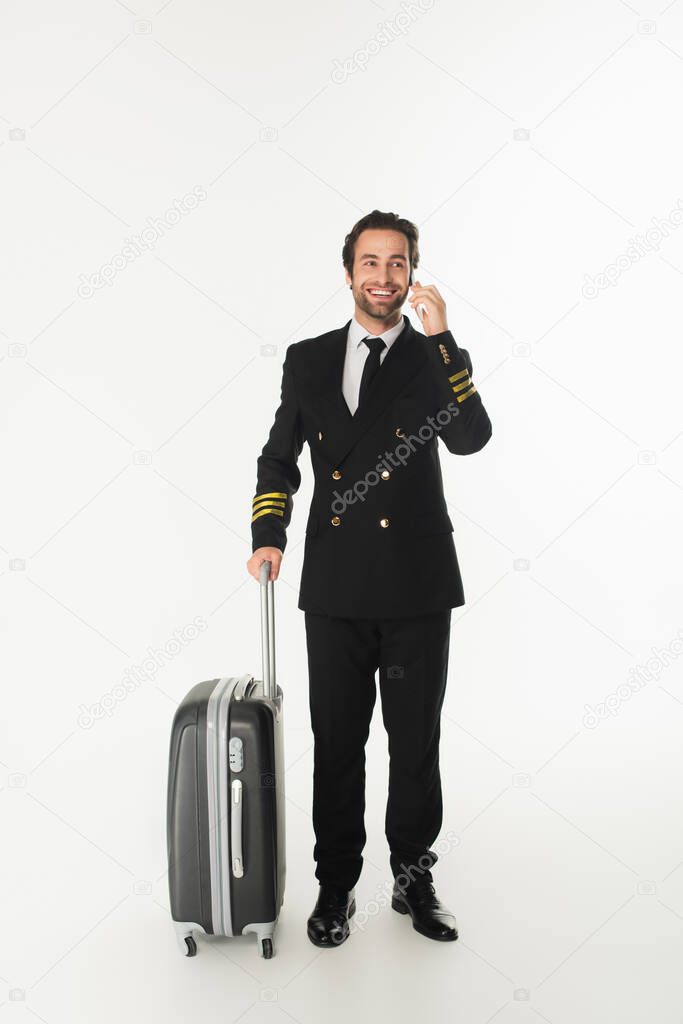 Smiling pilot talking on mobile phone near suitcase on white background 