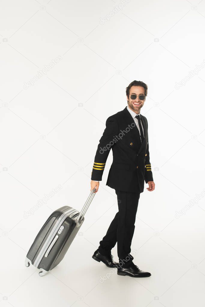 Smiling pilot in sunglasses holding suitcase while walking on white background 