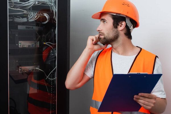 Side view of workman with clipboard talking on smartphone near switchboard 