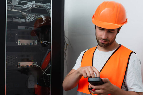 Workman in helmet holding electrical tester near switchboard 