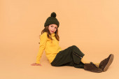teljes hosszúságú kilátás gyermek sárga garbó, kötött kalap és nadrág ül bézs