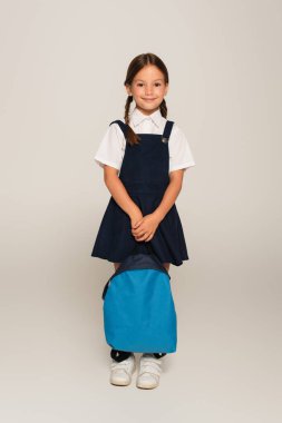 Gülümseyen okul çocuğunun gri üzerinde mavi sırt çantasıyla duruşu.