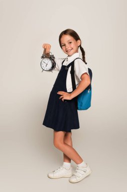 cheerful schoolgirl with blue backpack showing large alarm clock while standing with hand on hip on grey clipart
