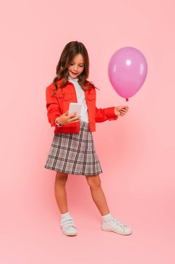 Moda elbiseli çocuk elinde cep telefonu ve pembe balonla duruyor.
