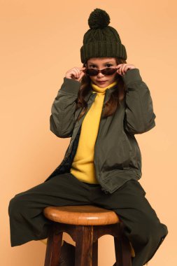 child in trendy autumn outfit looking at camera over stylish sunglasses isolated on beige clipart