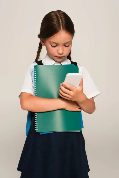 グレーで隔離された携帯電話を見ながらノートを持っている小学生は — ストック写真