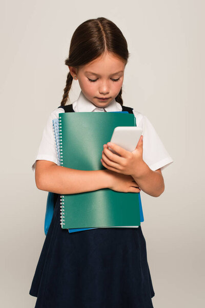 schoolchild holding notebooks while looking at mobile phone isolated on grey