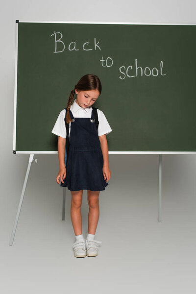 disappointed schoolgirl standing with bowed head near chalkboard with back to school lettering on grey