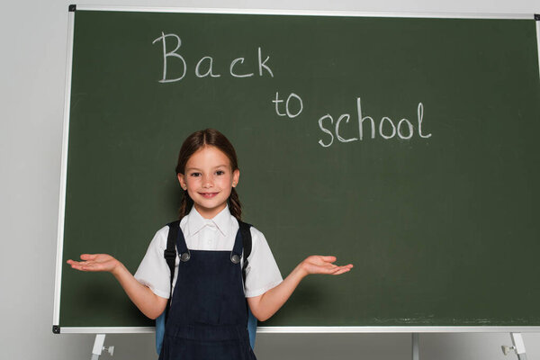 cheerful schoolgirl pointing with hands near chalkboard with back to school lettering on grey