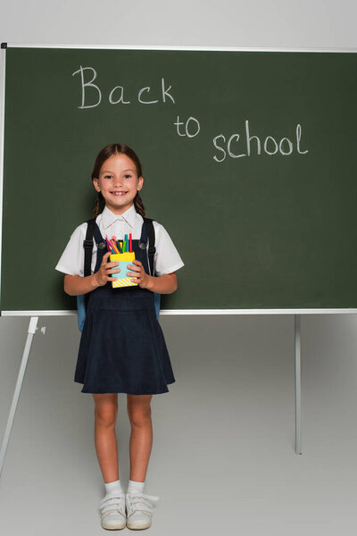 pleased schoolkid holding pen holder near chalkboard with back to school lettering on grey