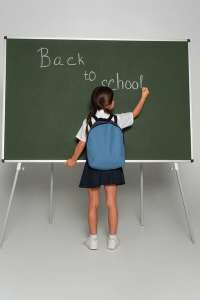 back view of schoolgirl with blue backpack writing back to school lettering on chalkboard on grey
