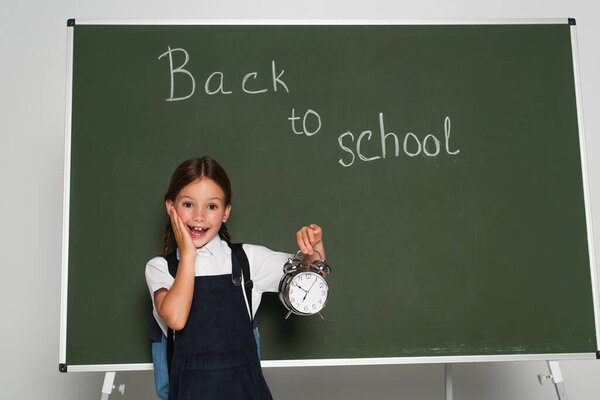 amazed schoolkid holding alarm clock near chalkboard with back to school lettering on grey